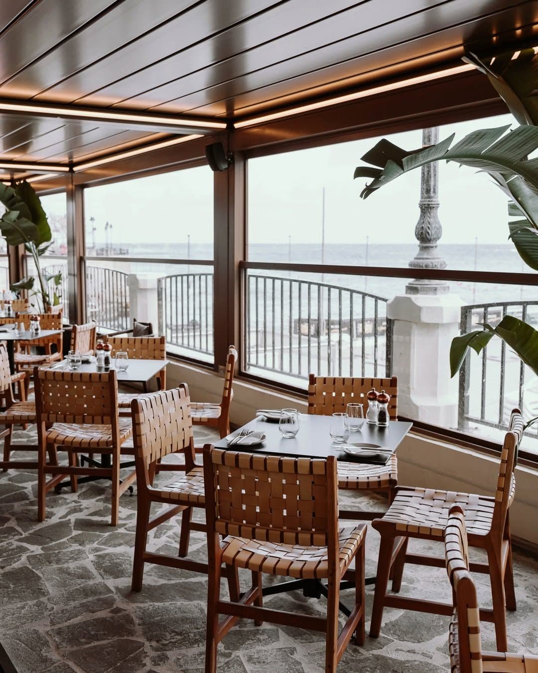 Wooden tables and woven chairs beside floor-to-ceiling windows facing the sea