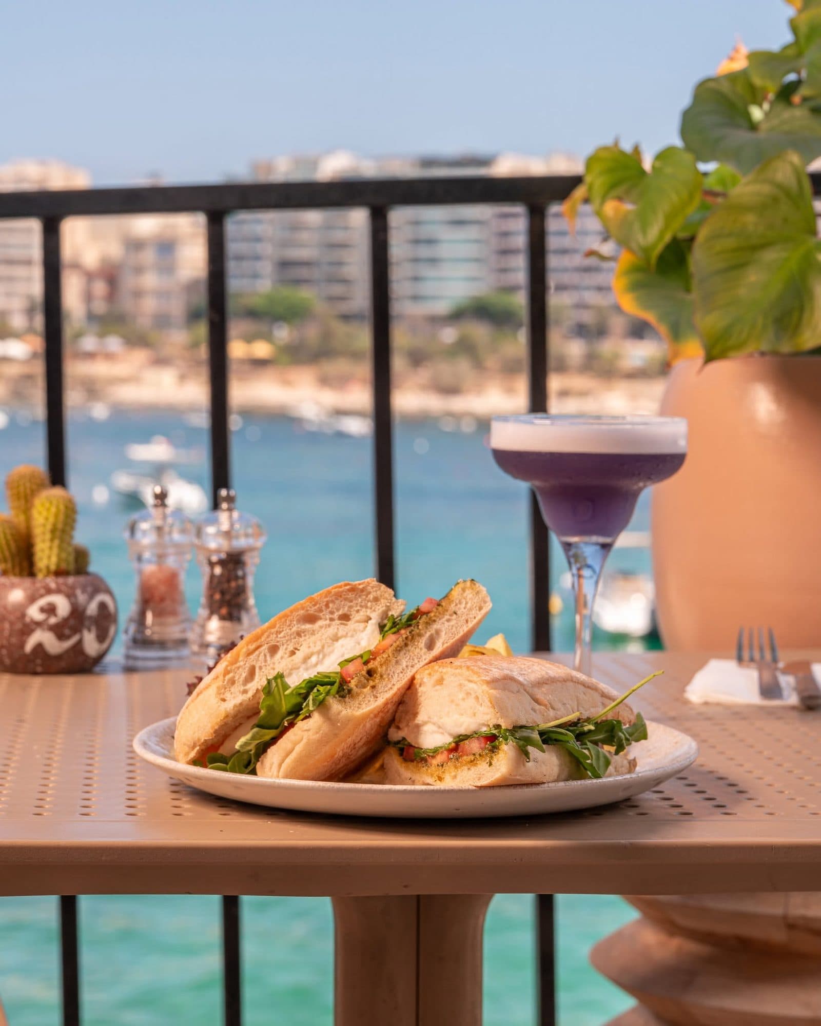 A ciabatta sandwich and a violet cocktail on the terrace with turquoise water behind