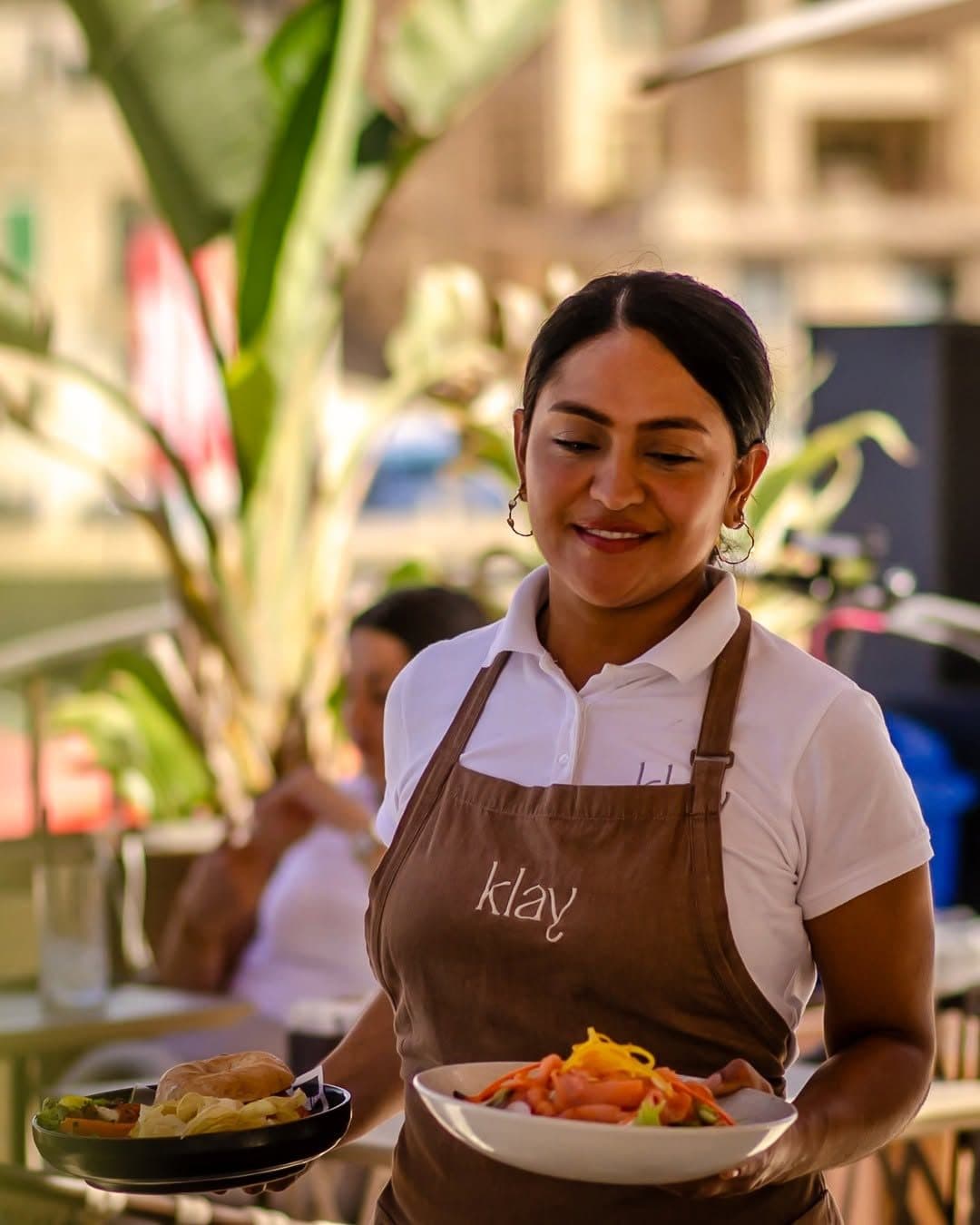 A Klay server in a branded apron carrying plates out to the terrace